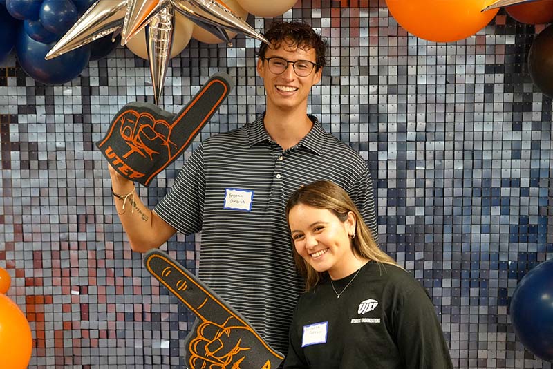 Two attendees pose together in front of a decorative balloon backdrop during a UTEP Engineering homecoming event, holding foam finger props and smiling for a commemorative photo.